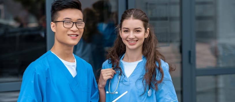A asian young man and caucasian young woman wearing blue scrubs and smiling at the camera 